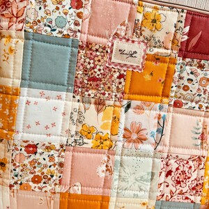 Op de afbeelding: Een patchwork etui met een verscheidenheid aan bloempatronen in roze, geel en oranje. De vierkanten zijn gewatteerd met een lichtbeige draad. Een klein label met de tekst "by Landyn" is op de voorkant genaaid.