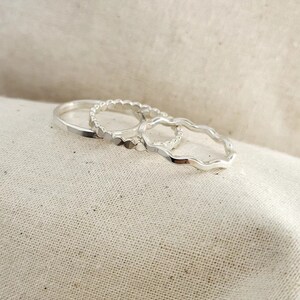 May include: Four silver rings are shown, each with a different design. One is a plain band, another has small beaded details, and a third has a wavy edge. The rings are set against a neutral, textured backdrop.