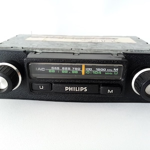 May include: A black Philips car radio with a silver knob on each side. The radio has a digital display with the text "IAC 545 625 750 100 1500 kHz M 88 92.96 101 104 MHz U" and the word "PHILIPS" printed on the front.