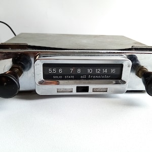 Könnte beinhalten: Ein vintage Silber-Autoradio mit schwarzen Knöpfen und einer silbernen Frontplatte. Das Radio hat ein Zifferblatt mit Zahlen von 5,5 bis 16 und den Text "SOLID STATE all transistor". Das Radio hat zwei Knöpfe mit den Beschriftungen "MUSIC" und "VOICE".