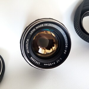May include: A black Minolta MC Rokkor-PF 55mm f/1.7 lens with a silver ring around the edge. The lens is made in Japan and has a black lens cap.
