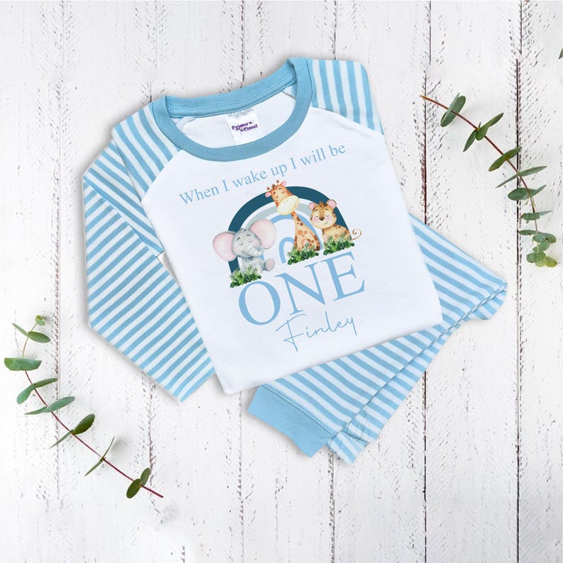 May include: Blue and white striped pajamas with a white long-sleeved shirt featuring a rainbow, elephant, giraffe, and tiger illustration. The shirt has the text "When I wake up I will be ONE Finley".