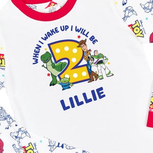 May include: A white long-sleeve shirt with red trim featuring a graphic of Toy Story characters, a large yellow number 2, and the text "WHEN I WAKE UP I WILL BE" and "LILLIE". The sleeves have a repeating pattern of Toy Story logos and characters.