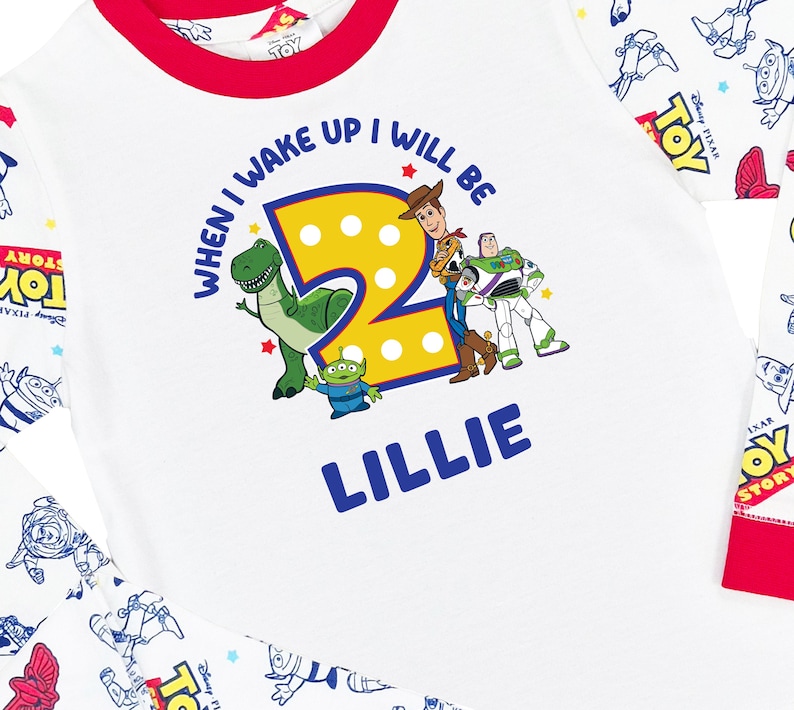 May include: A white long-sleeve shirt with red trim featuring a graphic of Toy Story characters, a large yellow number 2, and the text "WHEN I WAKE UP I WILL BE" and "LILLIE". The sleeves have a repeating pattern of Toy Story logos and characters.