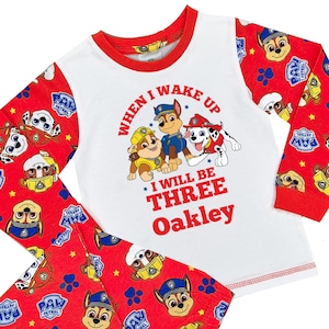 May include: A white and red Paw Patrol pajama set. The long-sleeved top features the text "When I wake up I will be Three Oakley" with Paw Patrol characters. The red pants and sleeves have a Paw Patrol pattern.