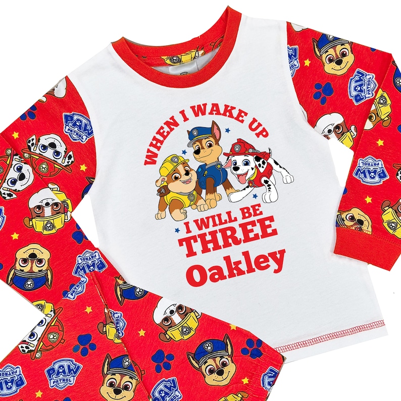 May include: A white and red Paw Patrol pajama set. The long-sleeved top features the text "When I wake up I will be Three Oakley" with Paw Patrol characters. The red pants and sleeves have a Paw Patrol pattern.