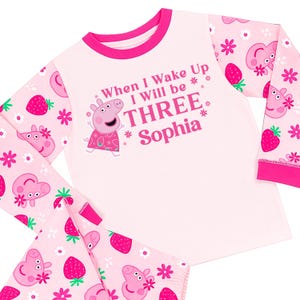 May include: A pink children's pyjama set with a Peppa Pig design and the text "When I Wake Up I Will be THREE Sophia". The long-sleeved top and trousers have a strawberry and floral pattern.