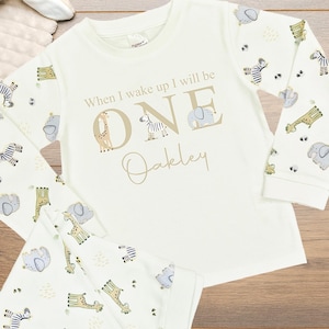 May include: Cream-coloured pyjama set with long sleeves and trousers. The sleeves and trousers are patterned with zebras, giraffes, and elephants. The shirt features the text "When I wake up I will be ONE" with a name below.