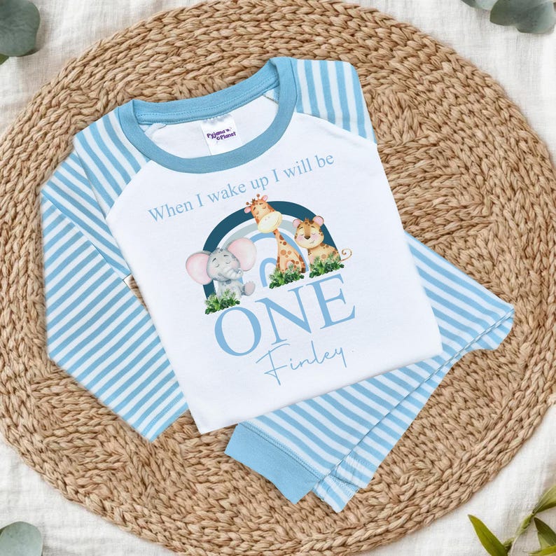 May include: Blue and white striped pajamas with a white long-sleeved shirt featuring a rainbow with animals and the text "When I wake up I will be ONE Finley".