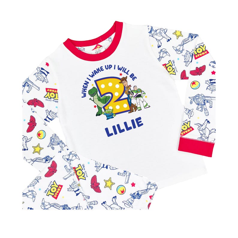 May include: A white long-sleeve pajama set with red trim featuring a Toy Story print. The shirt displays a large yellow number 2, characters Woody, Buzz Lightyear, Rex, and an alien, with the text "WHEN I WAKE UP I WILL BE 2" and "LILLIE".
