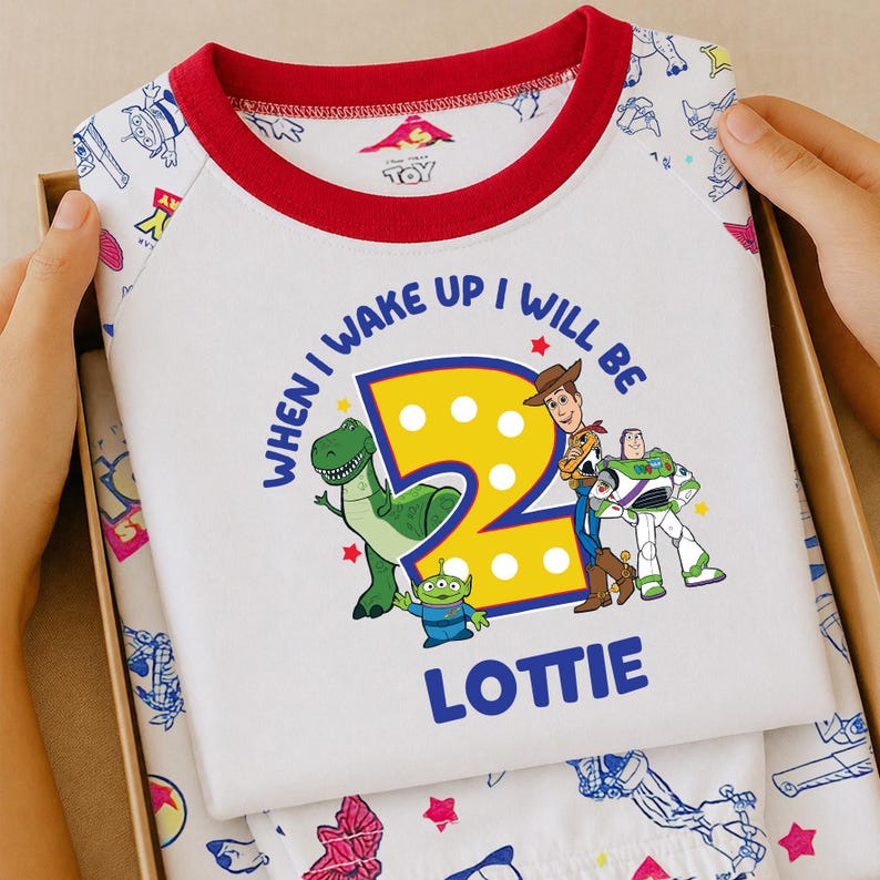 May include: A white t-shirt with a red crew neckline and raglan sleeves featuring a Toy Story graphic. The print includes a large yellow number two, Rex, an alien, Woody, and Buzz Lightyear, with the text "WHEN I WAKE UP I WILL BE 2 LOTTIE".