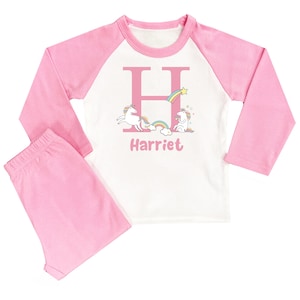 May include: Pink and white long-sleeved pyjamas with a unicorn and rainbow design. The pyjamas have a white top with a pink letter H and the name "Harriet" printed on it.