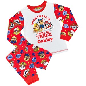 May include: A two-piece pajama set featuring a white top with a red trim and red sleeves. The top has a graphic of Paw Patrol characters and the text "WHEN I WAKE UP I WILL BE THREE Oakley". The red pants have a Paw Patrol pattern.