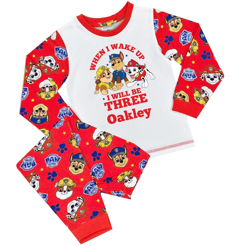 May include: A two-piece pajama set featuring a white top with a red trim and red sleeves. The top has a graphic of Paw Patrol characters and the text "WHEN I WAKE UP I WILL BE THREE Oakley". The red pants have a Paw Patrol pattern.