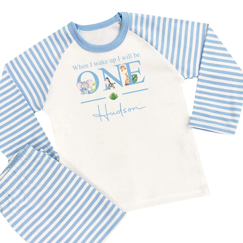 May include: A white and blue striped long-sleeved pajama set with a graphic of the word "ONE" with animal illustrations. The text "When I wake up I will be" is above the graphic and the name "Hudson" is below the graphic.