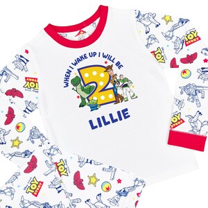 May include: A white long-sleeve shirt with red trim featuring a Toy Story graphic with the text "WHEN I WAKE UP I WILL BE 2" and the name "LILLIE" in blue. The sleeves and pants have an all-over print of Toy Story characters and logos.