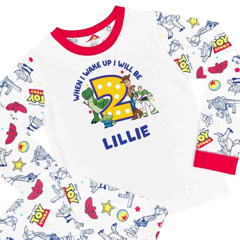 May include: A white long-sleeve shirt with red trim featuring a Toy Story graphic with the text "WHEN I WAKE UP I WILL BE 2" and the name "LILLIE" in blue. The sleeves and pants have an all-over print of Toy Story characters and logos.