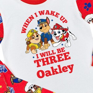 May include: White and red toddler's pajama set featuring a graphic of three cartoon dogs from Paw Patrol. The shirt reads "When I Wake Up I Will Be Three Oakley" in red. The sleeves and pants have a red background with Paw Patrol characters.