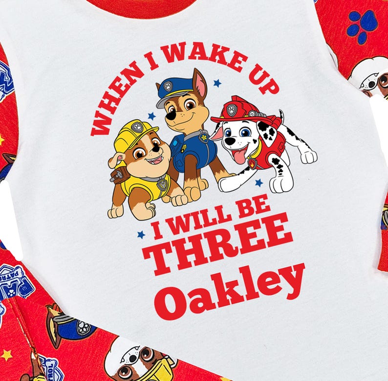 May include: White and red toddler's pajama set featuring a graphic of three cartoon dogs from Paw Patrol. The shirt reads "When I Wake Up I Will Be Three Oakley" in red. The sleeves and pants have a red background with Paw Patrol characters.