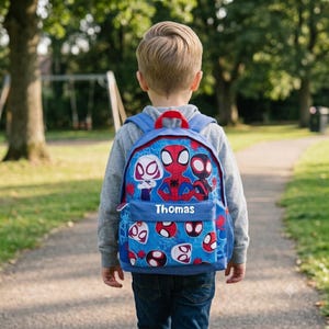 May include: A blue backpack featuring a repeating pattern of Spider-Man characters and masks. The front pocket displays the name Thomas in white lettering. The bag has a red top handle, blue shoulder straps, and a zippered main compartment.