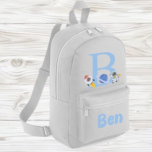 May include: A light grey backpack with a front zipped pocket and adjustable shoulder straps. The backpack features a large blue letter "B" with a rocket ship, planet, and astronaut graphic. The name "Ben" is printed in blue on the front pocket.