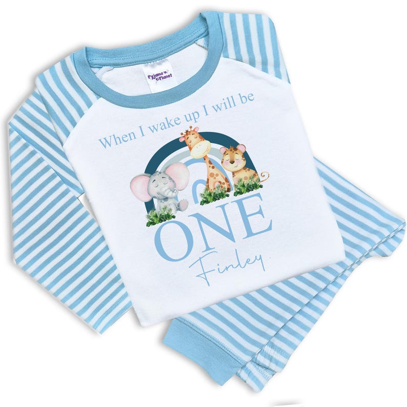 May include: Blue and white striped pajamas with a white long-sleeved shirt featuring a rainbow with a giraffe, elephant, and lion. The shirt also has the text "When I wake up I will be ONE Finley".