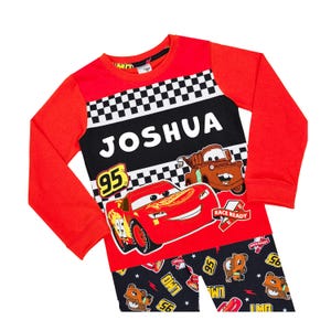 May include: A long-sleeve shirt and trousers set featuring a red and black colour scheme. The shirt displays the name JOSHUA, a checkered pattern, and graphics of Lightning McQueen and Mater from Cars, with the number 95 and the text RACE READY.