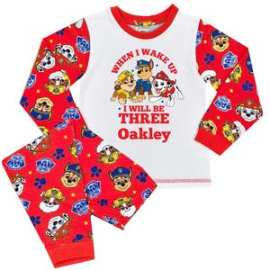 May include: A two-piece pajama set featuring a white top with red trim and sleeves. The top displays Paw Patrol characters and the text "When I Wake Up I Will Be Three Oakley." The red pants have a Paw Patrol pattern.