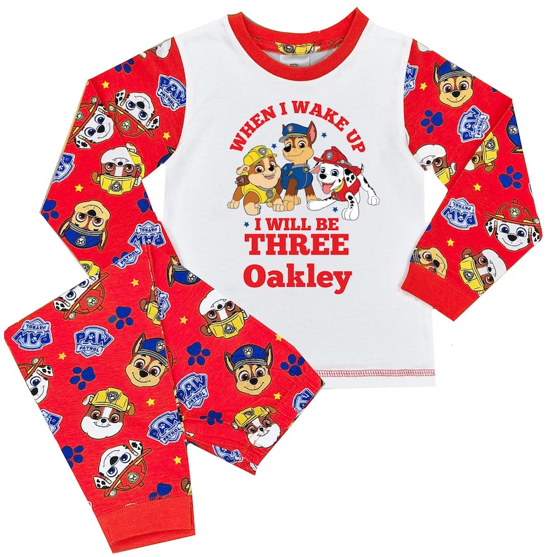 May include: A two-piece pajama set featuring a white top with red trim and sleeves. The top displays Paw Patrol characters and the text "When I Wake Up I Will Be Three Oakley." The red pants have a Paw Patrol pattern.
