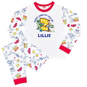 May include: White long-sleeve pajama set with red trim featuring a Toy Story print. The shirt displays the text "WHEN I WAKE UP I WILL BE 2" above a large yellow number two, Toy Story characters, and the name "LILLIE" in blue lettering.