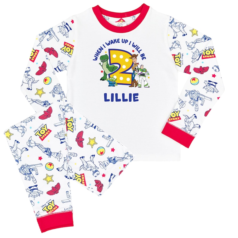 May include: White long-sleeve pajama set with red trim featuring a Toy Story print. The shirt displays the text "WHEN I WAKE UP I WILL BE 2" above a large yellow number two, Toy Story characters, and the name "LILLIE" in blue lettering.