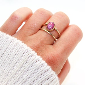 Könnte beinhalten: Ein goldener Ring mit einem rosafarbenen ovalen Edelstein in der Mitte. Der Ring wird am Zeigefinger einer Hand getragen.