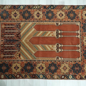 May include: A traditional Turkish prayer rug with a red background and intricate geometric patterns in shades of blue, green, and beige. The rug features a central design of four arches, symbolizing the four pillars of Islam.
