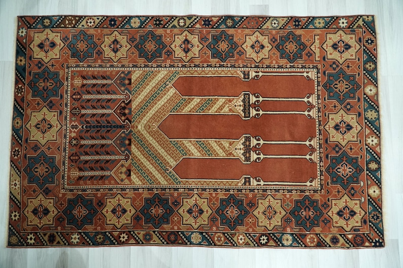 May include: A traditional Turkish prayer rug with a red background and intricate geometric patterns in shades of blue, green, and beige. The rug features a central design of four arches, symbolizing the four pillars of Islam.