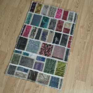 Può includere: Un tappeto patchwork rettangolare con una varietà di colori e motivi. Il tappeto è composto da pezzi rettangolari e quadrati in tonalità di verde, rosa, blu, grigio e viola. Il tappeto è posizionato su un pavimento in legno marrone chiaro.