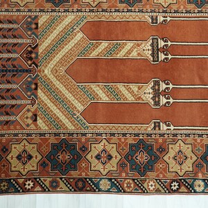 May include: A traditional prayer rug with a red background and intricate geometric patterns in shades of blue, beige, and brown. The rug features a central design of three arches, symbolizing the mihrab, a niche in a mosque that indicates the direction of Mecca.