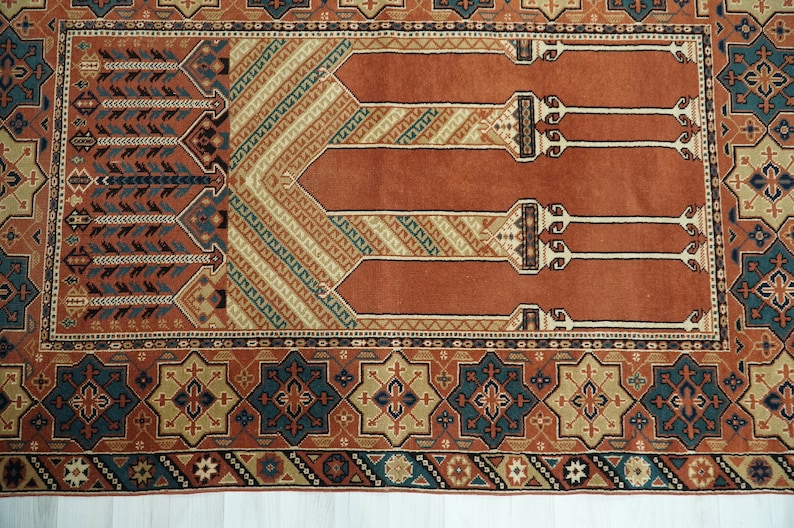 May include: A traditional prayer rug with a red background and intricate geometric patterns in shades of blue, beige, and brown. The rug features a central design of three arches, symbolizing the mihrab, a niche in a mosque that indicates the direction of Mecca.