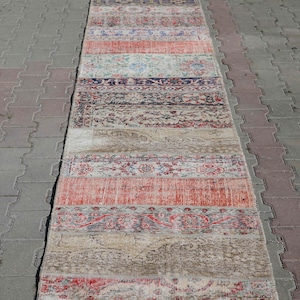 May include: A long, rectangular runner rug with a patchwork design. The rug features various rectangular sections in shades of red, beige, blue, and grey. The rug is placed on a grey brick surface.