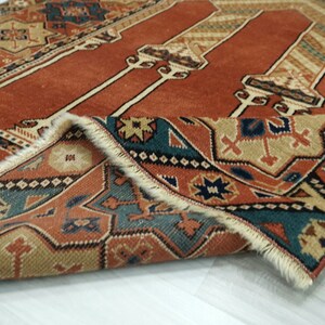 May include: A close-up of a folded rug with a red, orange, and blue geometric pattern. The rug has a fringe border and is made of wool.
