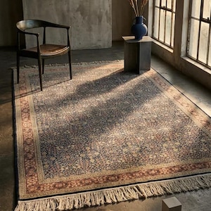 Könnte beinhalten: Ein rechteckiger Teppich mit detailliertem Blumenmuster in Marine-, Beige- und Rottönen. Der Teppich hat einen Fransenrand und liegt auf einem Betonboden. Das Bild zeigt auch einen Holzstuhl, eine blaue Vase und Bücher.