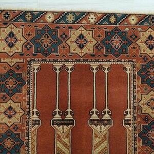May include: A traditional prayer rug with a red background and intricate geometric patterns in shades of blue, green, and beige. The rug features a central design with four columns and a border with repeating motifs.