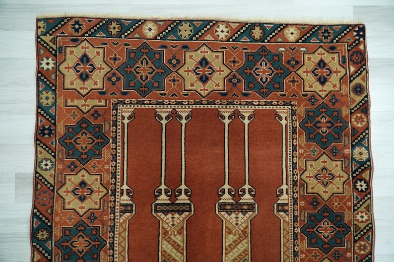May include: A traditional prayer rug with a red background and intricate geometric patterns in shades of blue, green, and beige. The rug features a central design with four columns and a border with repeating motifs.