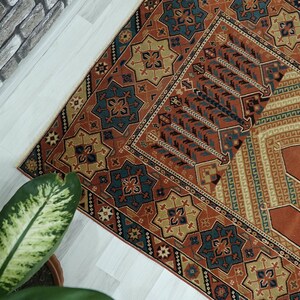 May include: A traditional prayer rug with a geometric pattern in shades of red, orange, blue, and beige. The rug has a central rectangular area with a repeating pattern of stylized trees.