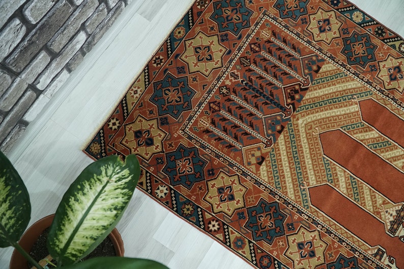May include: A traditional prayer rug with a geometric pattern in shades of red, orange, blue, and beige. The rug has a central rectangular area with a repeating pattern of stylized trees.