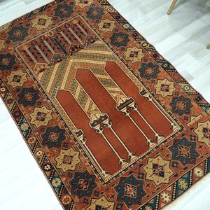 May include: A traditional prayer rug with a red center and intricate geometric patterns in shades of blue, green, and gold. The rug has a fringed border and is perfect for use in a home or mosque.