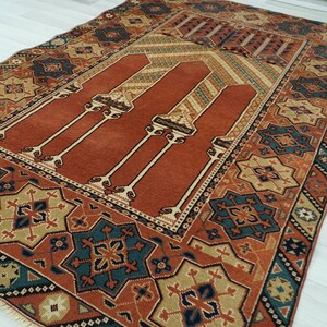 May include: A traditional Turkish prayer rug with a red center and intricate geometric patterns in shades of blue, green, and beige. The rug has a fringed edge and is made of wool.