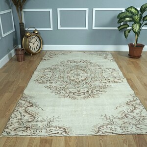 May include: Cream-colored, rectangular rug with an intricate brown floral pattern. The rug features a central medallion design and decorative borders. The rug is placed on a hardwood floor, adding warmth to the room.