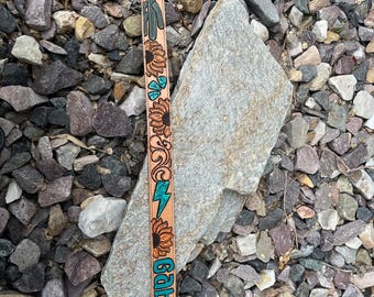 May include: A light brown leather strap with a floral and desert-themed design. The strap features sunflowers, a cactus, and turquoise accents. The name "Gabby" is written in turquoise letters. The strap is likely for a guitar or bag.