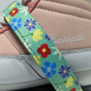 May include: A pair of pink and white sneakers with a floral patterned strap. The strap is green with yellow, red, blue, and purple flowers.
