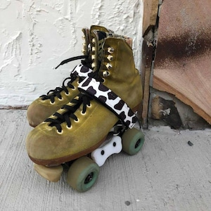 May include: A pair of green suede roller skates with black laces and a white and black cow print strap. The skates have white plastic wheels and a white plastic toe stop.